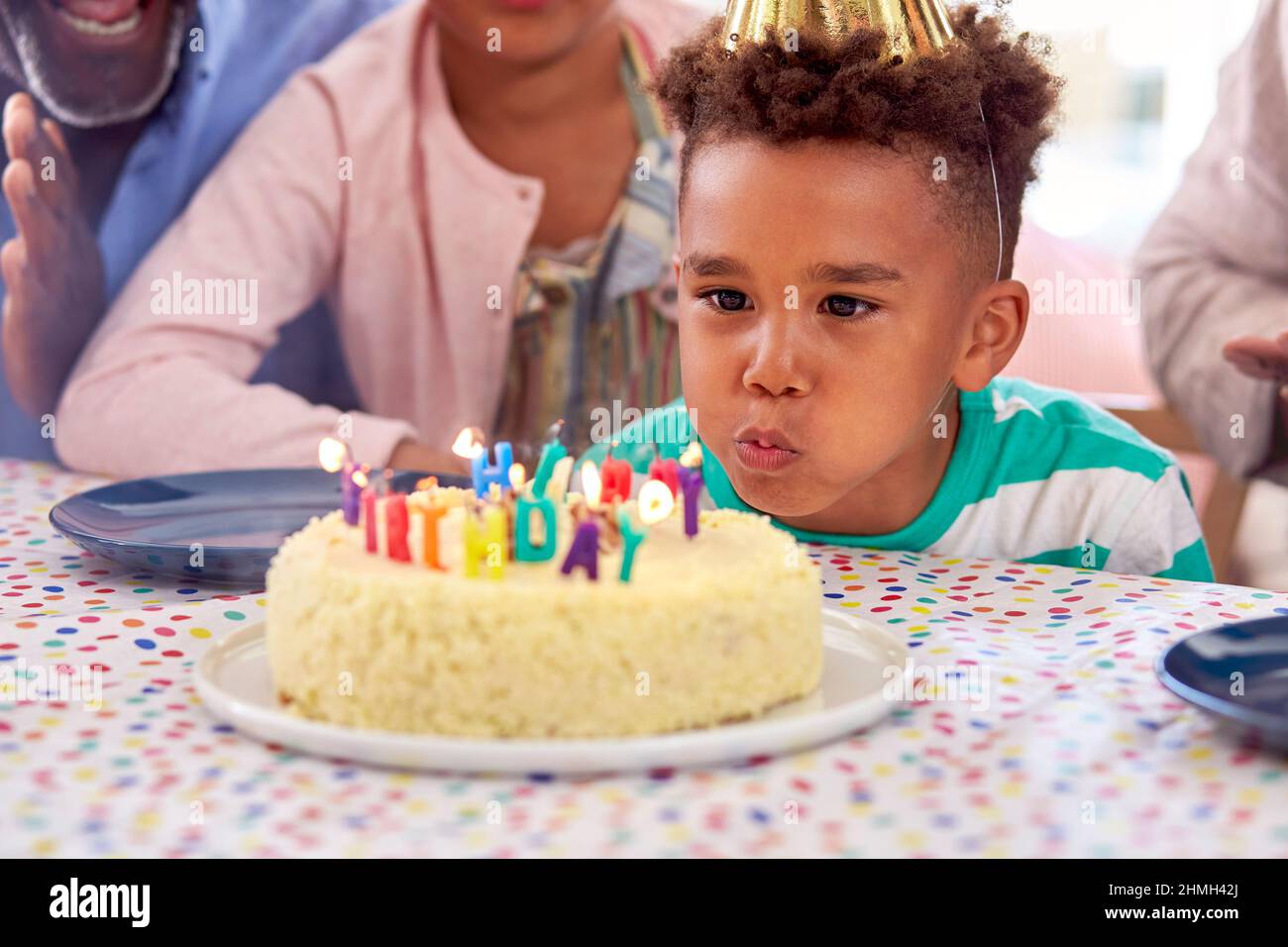 La famiglia che si siede intorno al tavolo a casa festeggia come il ragazzo soffia fuori le candele sulla torta di compleanno al partito Foto Stock