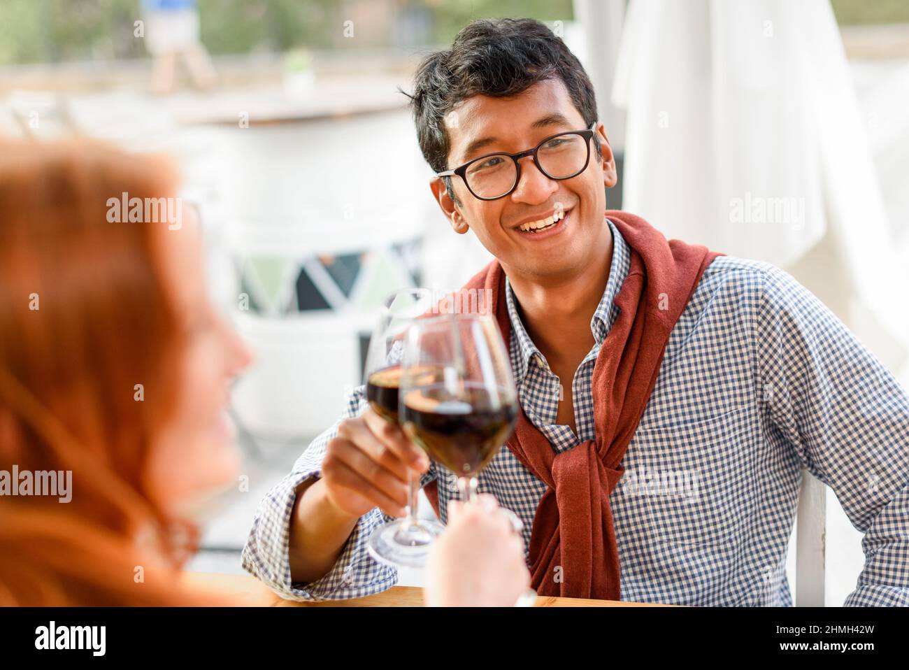 Positivo maschio etnico cliching bicchiere di vino rosso con un amico femmina e guardando via al tavolo in ristorante strada Foto Stock