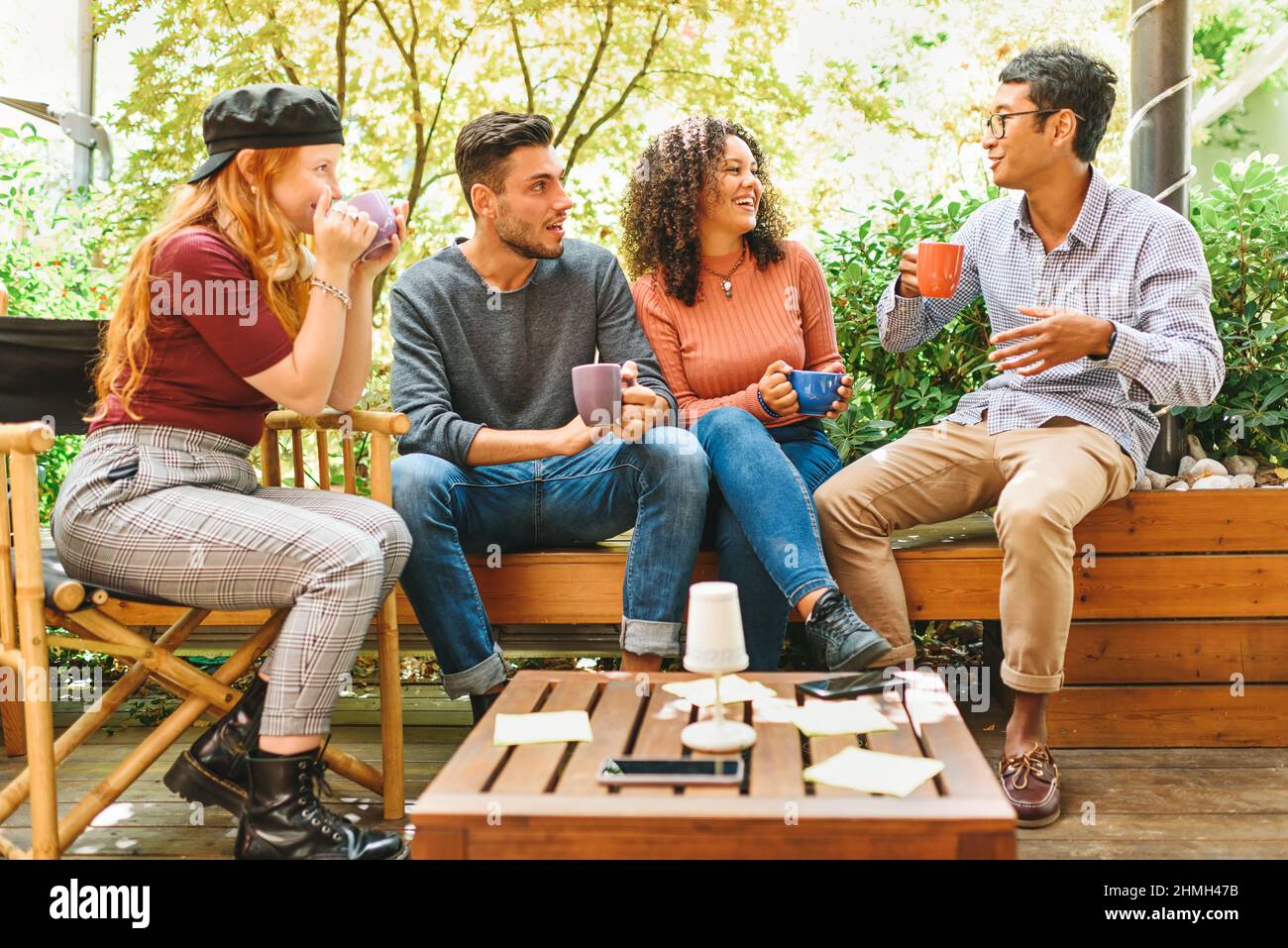 Gruppo di giovani amici multiculturali che si aggrappano su un patio che si agghiacciano insieme su tazze di caffè che ridono e scherzano su un alto sfondo chiave Foto Stock
