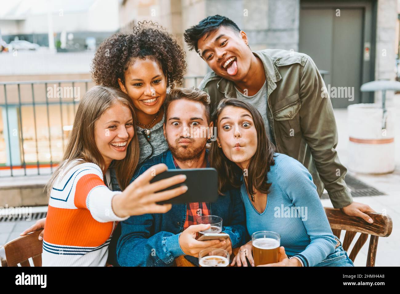 Gruppo di amici multirazziali positivi che fanno volti divertenti mentre si prende il ritratto di auto sullo smartphone al tavolo del caffè Foto Stock