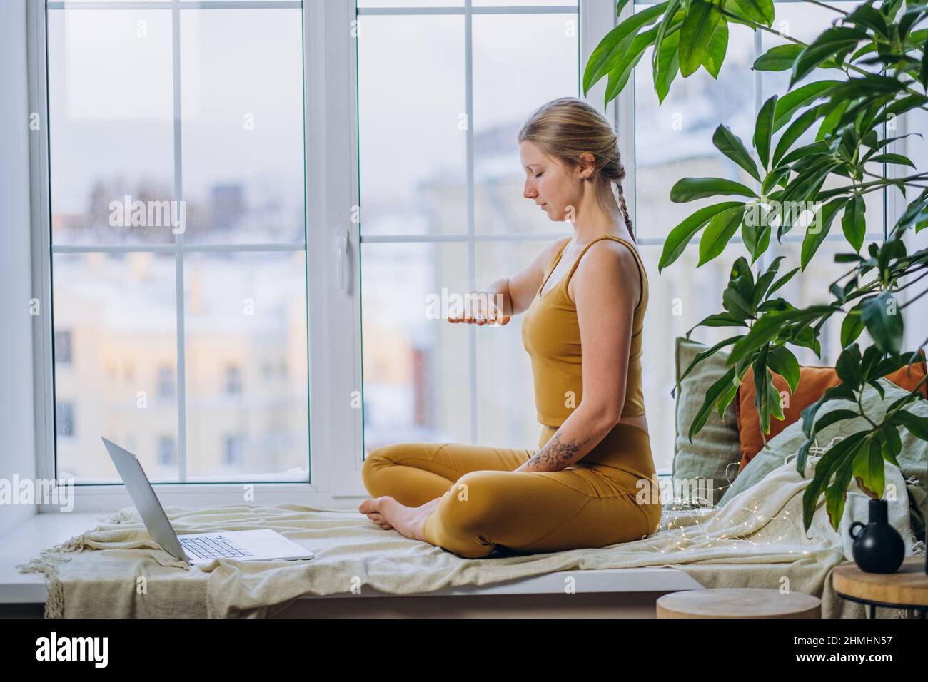 Giovane donna bionda in cima gialla e leggings medita in posa yoga seduta sul davanzale coperto di coperta di coperta e cuscini utilizzando un computer portatile Foto Stock
