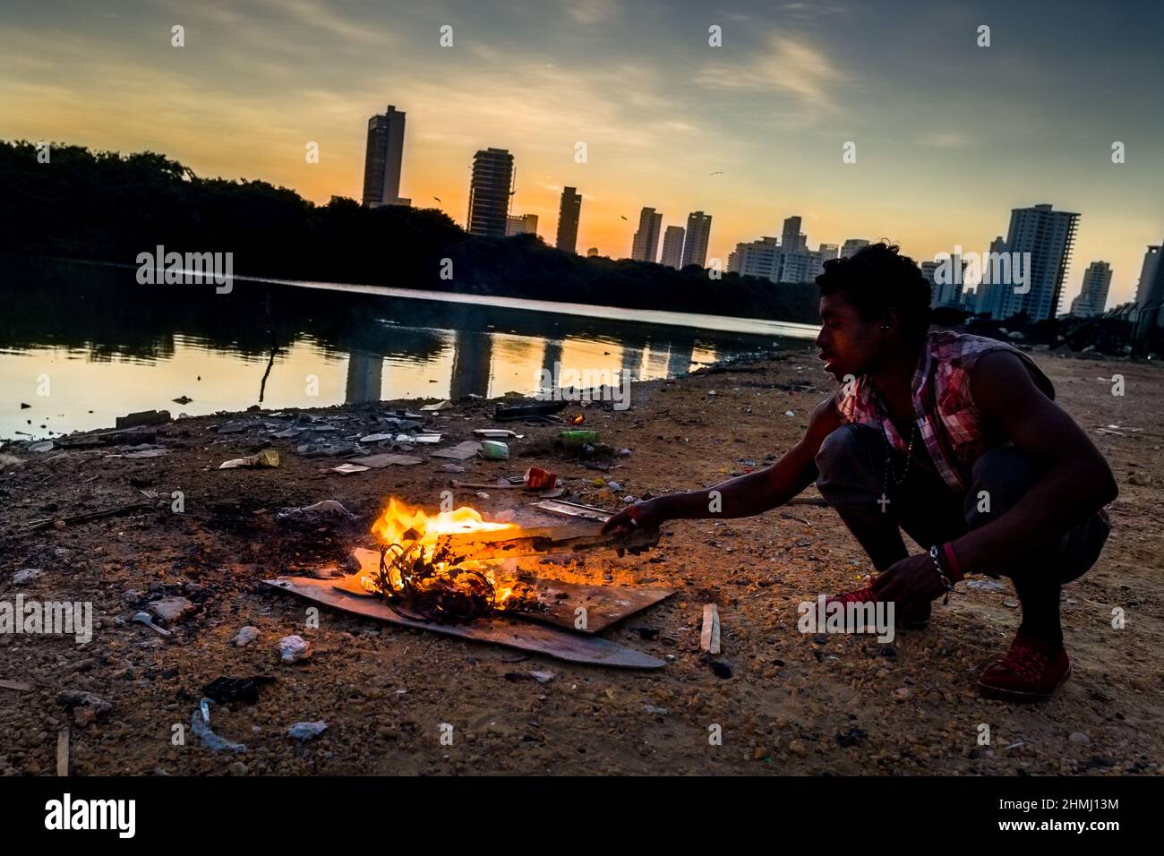Un giovane uomo afro-colombiano brucia fili elettrici per recuperare il rame sulla riva della laguna marina a Bahía de Manga, un lussuoso quartiere di carta Foto Stock