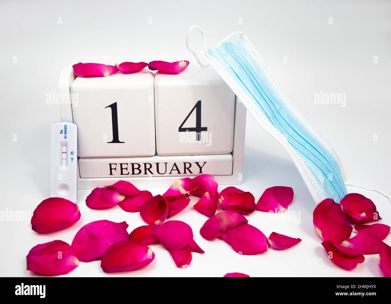 Febbraio 14 calendario in legno bianco su sfondo bianco con petali di rosa rossa. Accanto a lui c'è una maschera medica e un test positivo covid Foto Stock