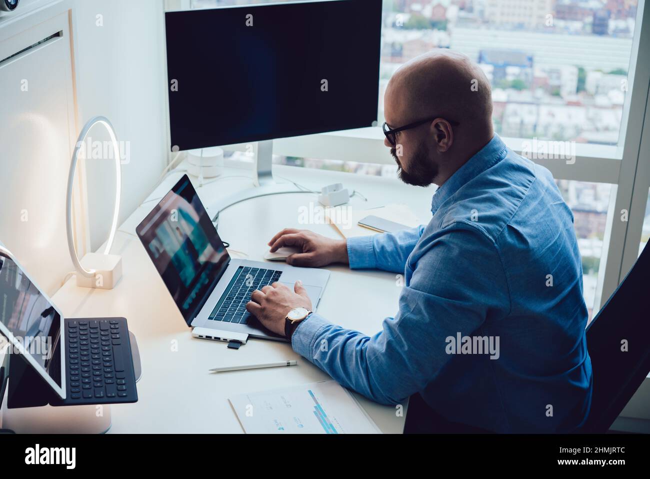 Un dipendente di sesso maschile serio che digita su netbook seduto a un tavolo con computer e tablet Foto Stock