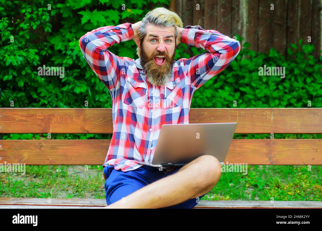 Uomo sorridente con un computer portatile seduto su una panca nel parco. Buon maschio con il computer. Gioco online. Foto Stock
