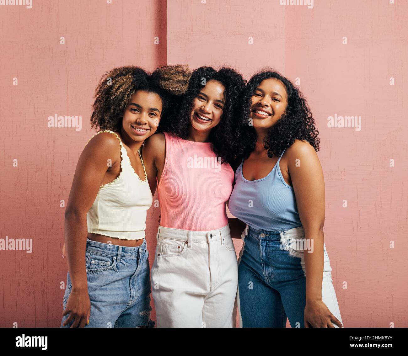 Tre donne positive con capelli ricci in piedi insieme contro una parete rosa e guardando la fotocamera Foto Stock