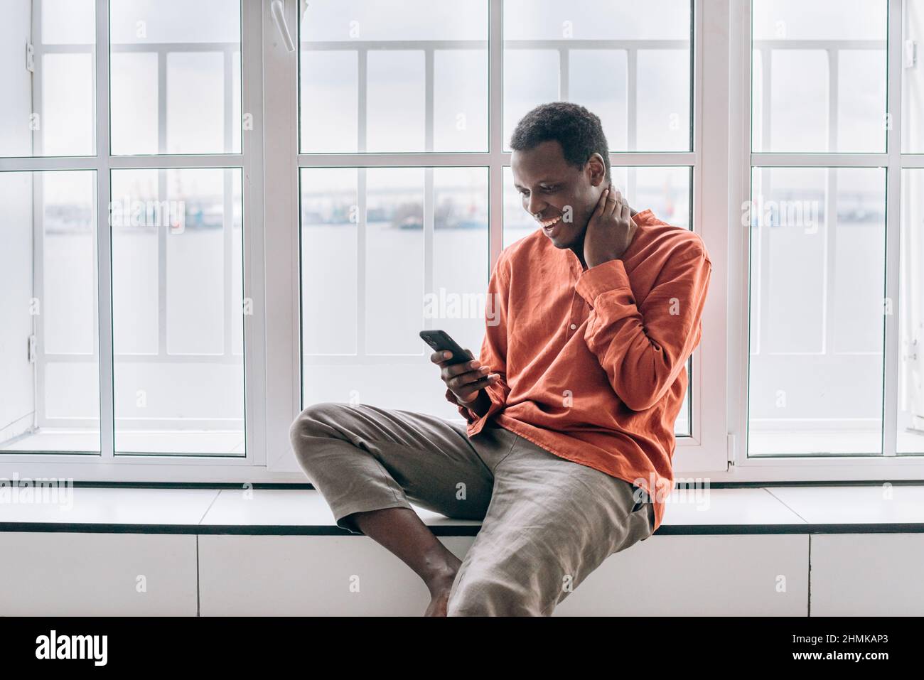 Il ragazzo afro-americano positivo in elegante camicia arancione guarda dalla finestra sorridendo con il telefono cellulare moderno nella stanza luminosa di casa Foto Stock