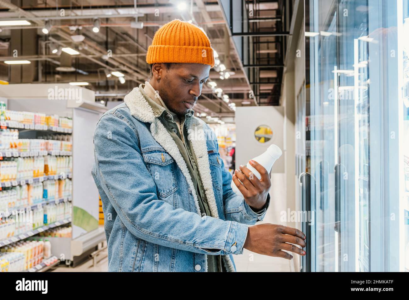 Il ragazzo afro-americano positivo in giacca di denim e cappello arancione prende la bottiglia di latte dalla scatola del frigorifero in vista laterale del moderno supermercato Foto Stock