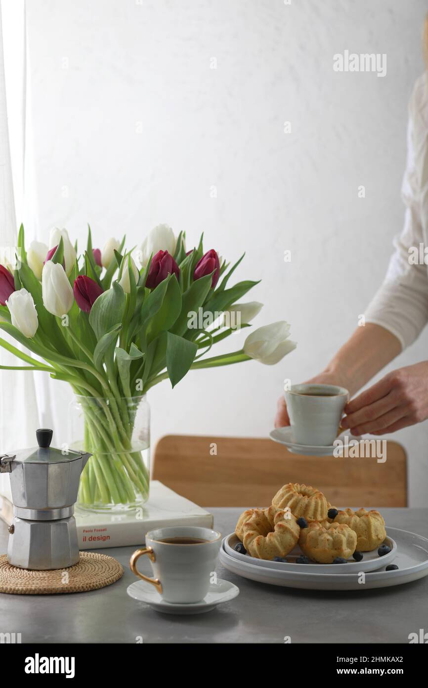 Mini panetteria con frutta sul piatto, due tazze di caffè e fiori di tulipano banch in un vaso. Ragazza che tiene una tazza di caffè. Foto Stock