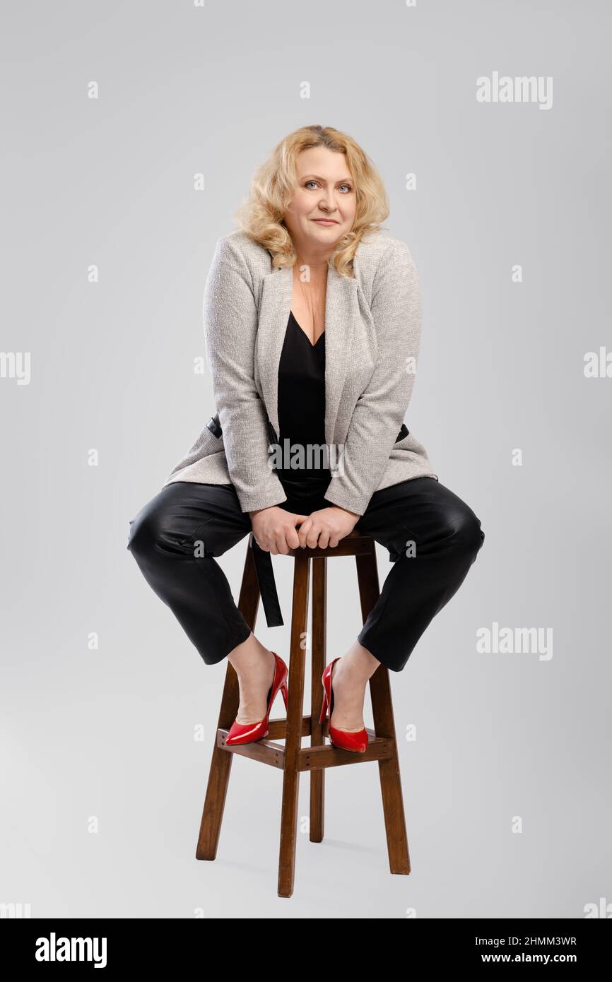 Ritratto di donna anziana in pantaloni di pelle e blazer seduta su sedia su  sfondo grigio studio Foto stock - Alamy