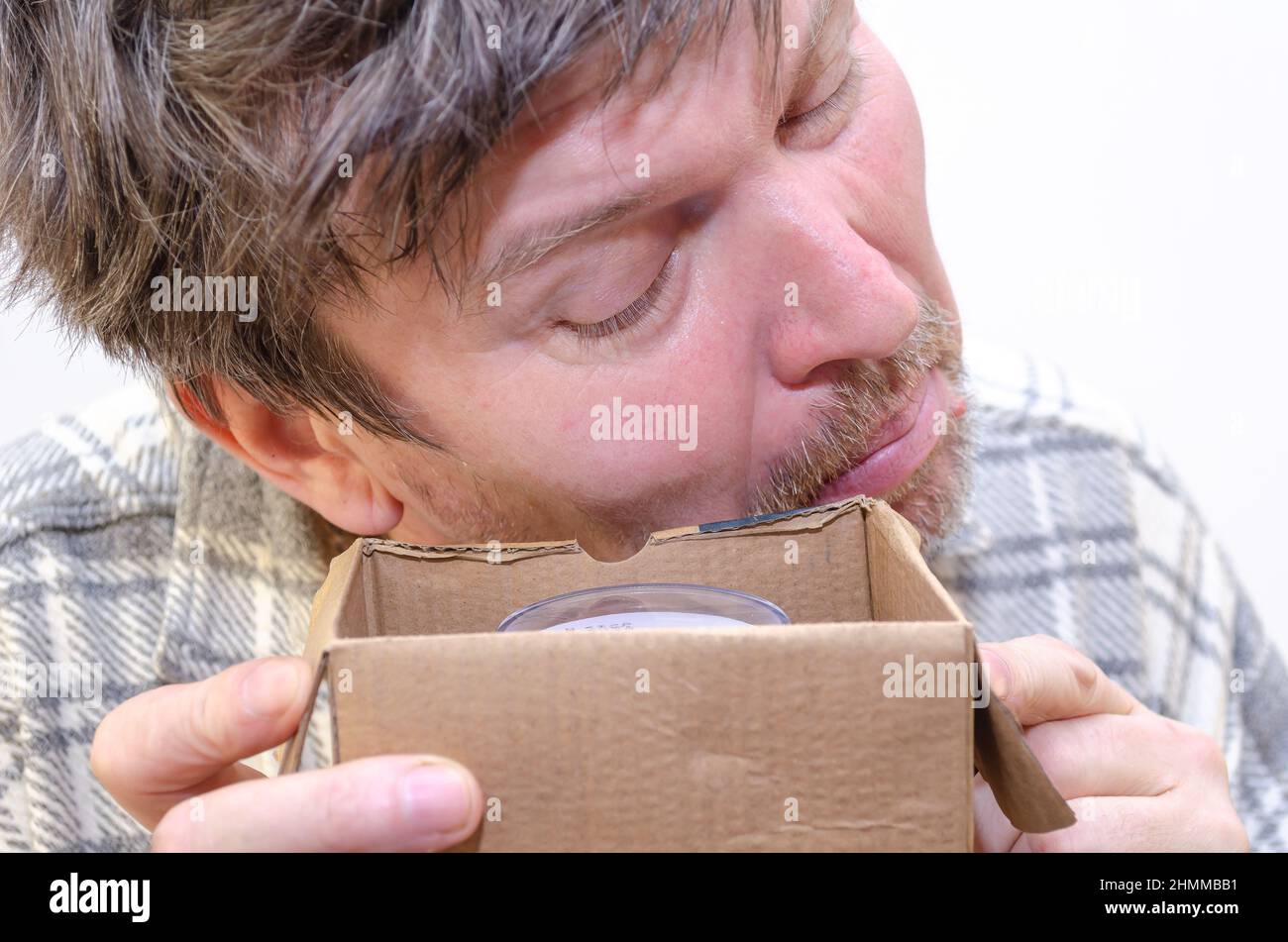 Un uomo adulto guarda con cautela in una scatola di cartone aperta. Un nuovo contatore dell'acqua si trova in una piccola scatola. Un maschio in una camicia a quadri è contro il wal grigio chiaro Foto Stock