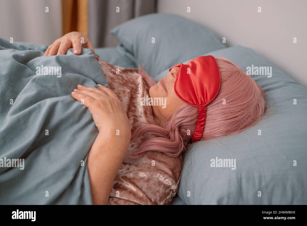 Positivo rilassato 50s donna sdraiata a letto con maschera per dormire su un letto grigio durante il fine settimana pigro Foto Stock