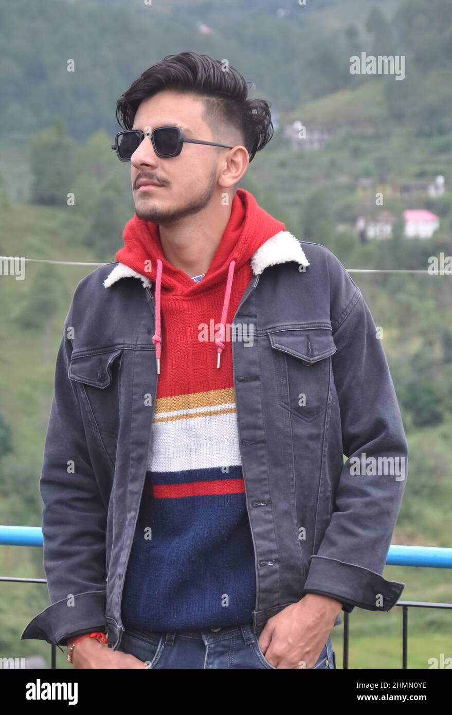 Un giovane indiano affascinante che indossa occhiali da sole in posa con le mani in tasca, in piedi all'aperto con lo sguardo lontano Foto Stock