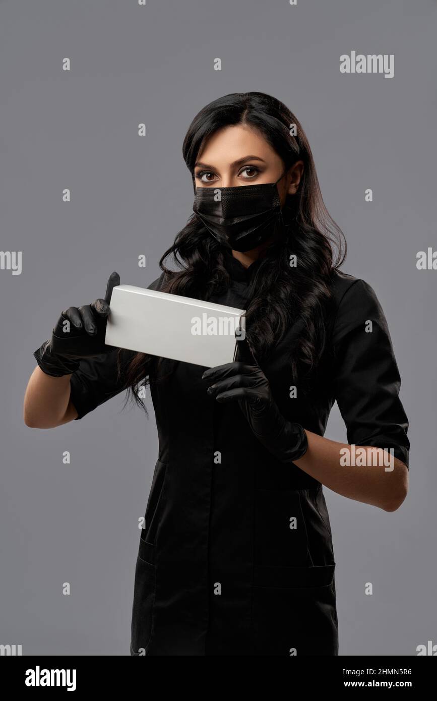 Medico donna in uniforme nera, maschera viso e guanti che tengono in mano la scatola cosmetica mentre si posa su sfondo grigio studio. Concetto di cura della pelle e cosmetologia. Foto Stock