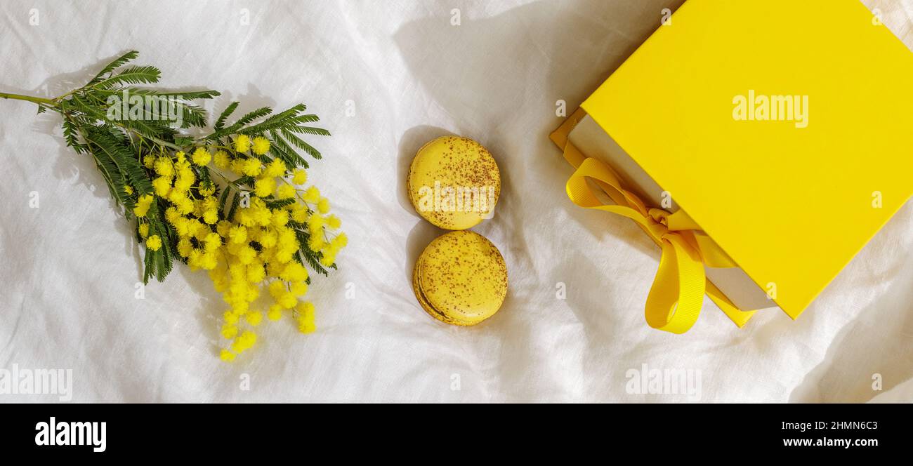 Rami di fiori gialli di mimosa su lenzuola di lino bianco, maccheroni gialli francesi e scatola regalo gialla. Spazio copia per Complimenti. Saluto per Foto Stock