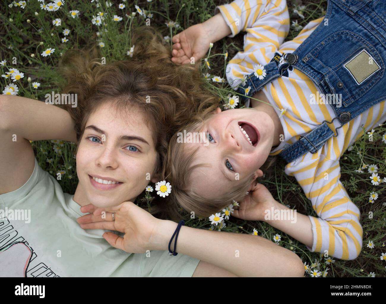 Il fratello più giovane e la sorella più anziana giacciono sull'erba tra le margherite con un ampio sorriso e una sensazione di felicità. Umore estivo, spensierata e gioiosa infanzia. Lui Foto Stock