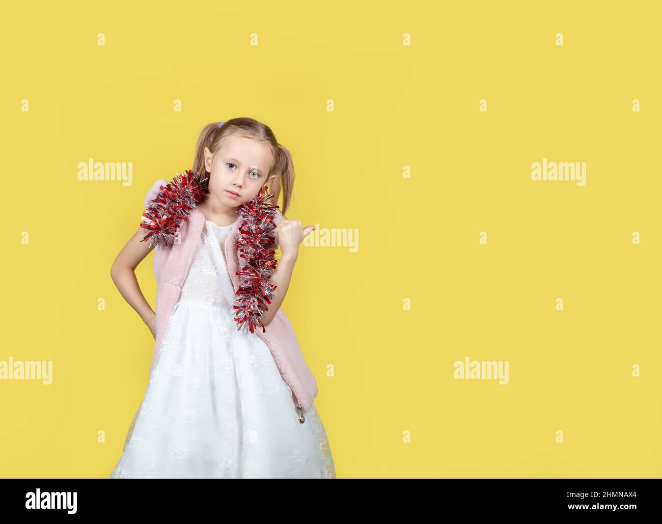 Ritratto di bambina carina in un abito elegante e con tinsel facendo un gesto di puntamento con posto per il testo su sfondo giallo. Il concetto Foto Stock