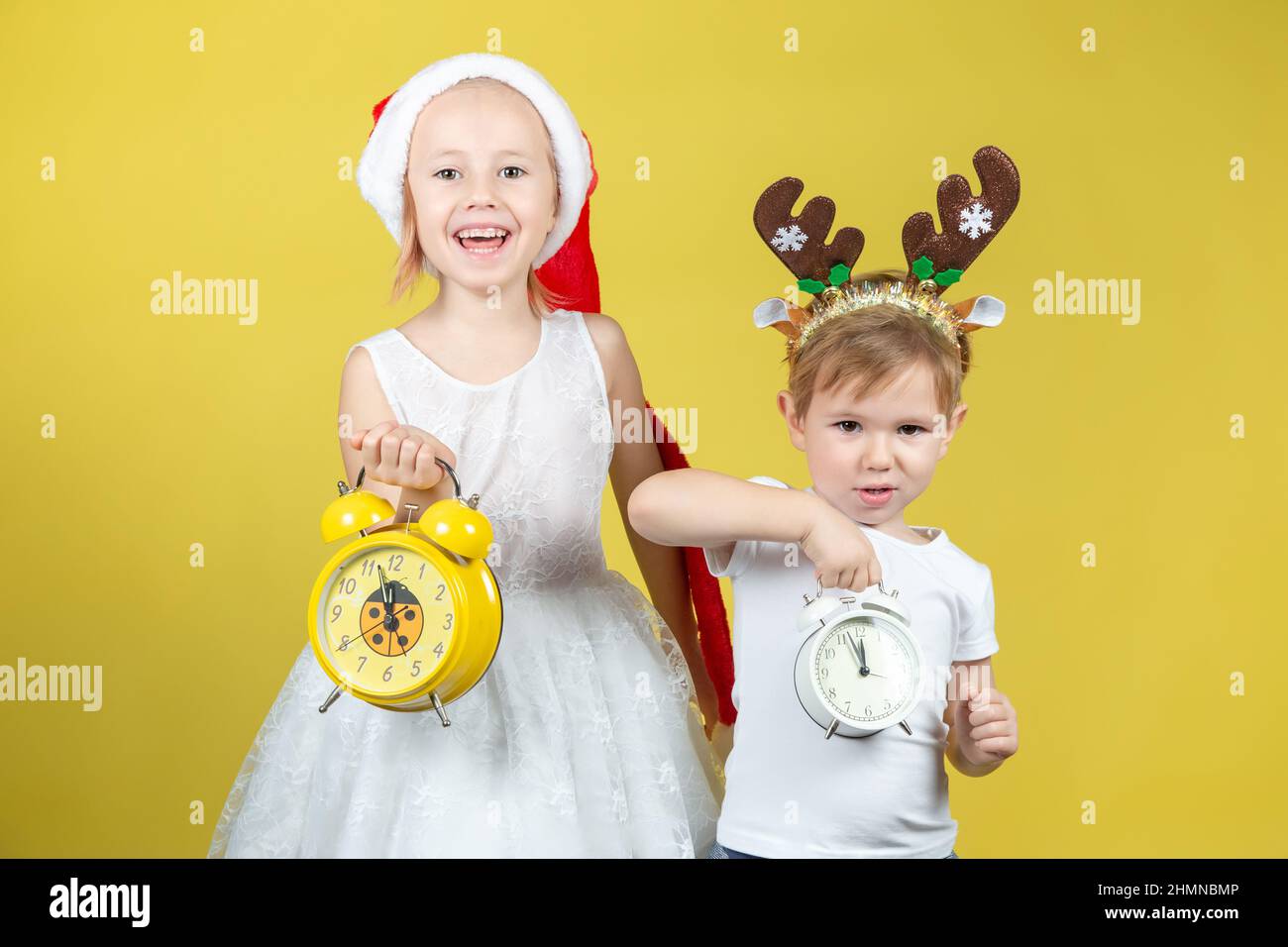 Adorabili bambini caucasici a natale a Santa Hat con due sveglia in mani. Un luogo per il testo. Studio girato su sfondo giallo. La conce Foto Stock