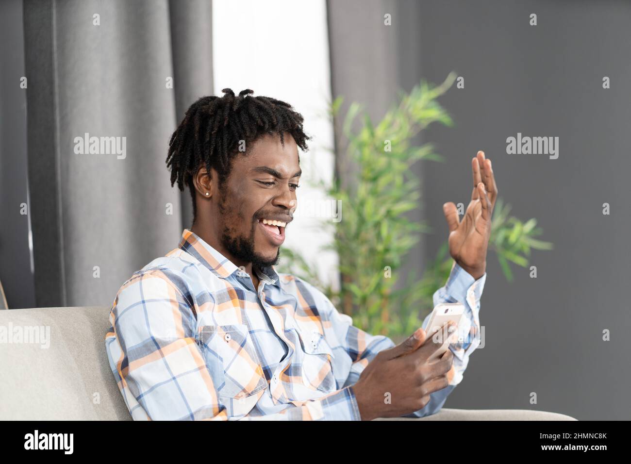 Felice ridere studente afroamericano utilizzare smartphone per videochiamare o guardare video online. Ritratto di uomo felice gesturing guardando lo smartphone seduto sul divano. Concetto di social media. Foto Stock