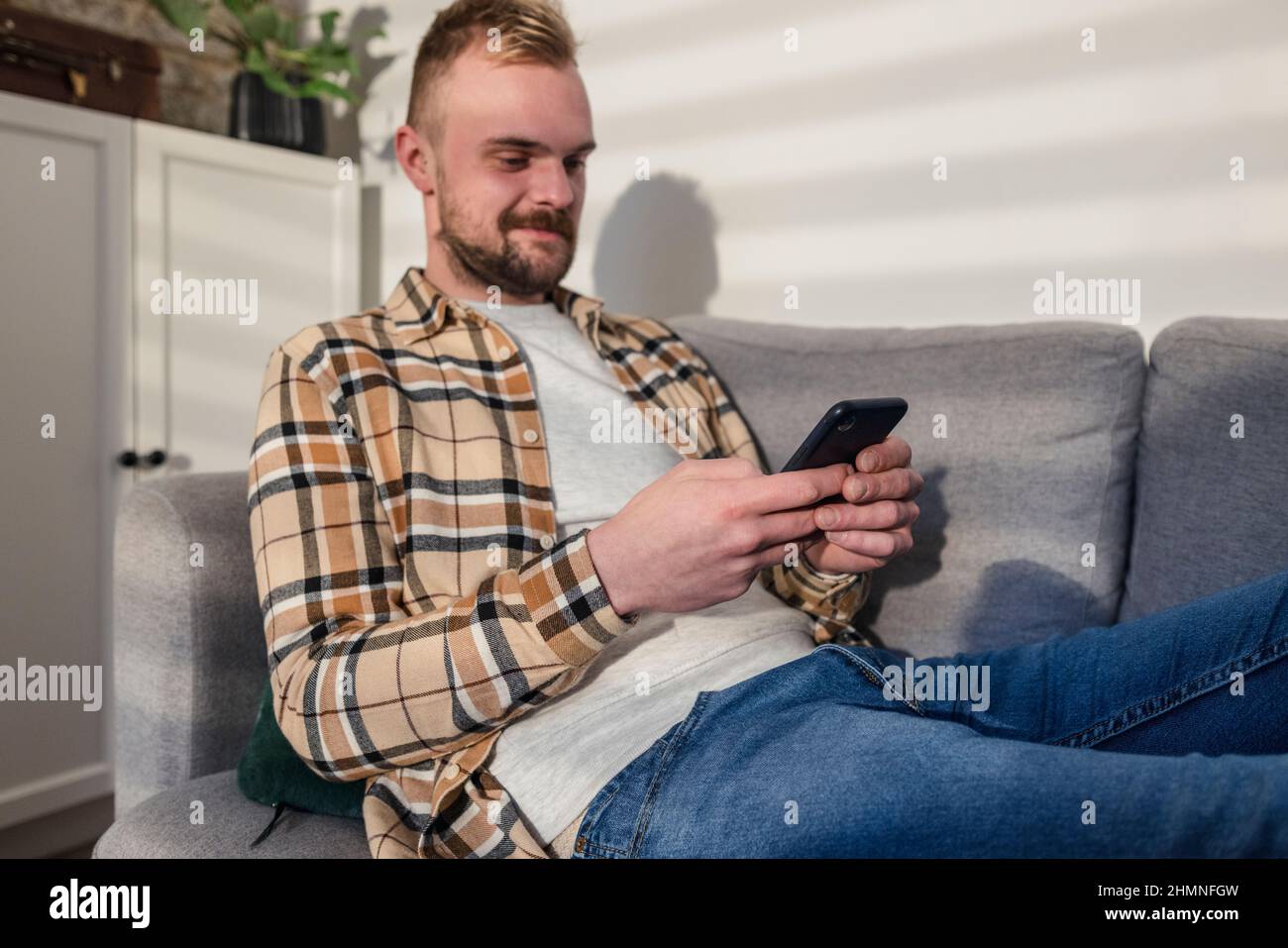 Uno scatto laterale di un giovane uomo che si trova sul divano nel suo soggiorno rilassante, sta usando il suo smartphone. Foto Stock