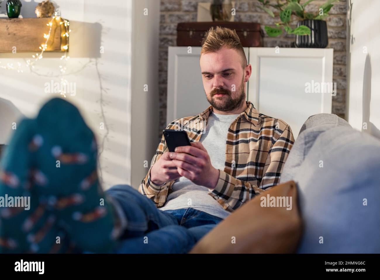 Uno scatto in vista frontale di un giovane uomo sdraiato sul divano nel suo soggiorno rilassante, sta usando il suo smartphone. Foto Stock