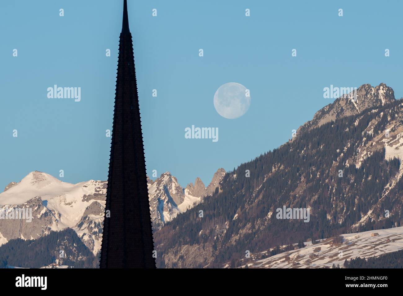 Schaan, Liechtenstein, 19 gennaio 2022 Luna piena scenario al mattino dietro la torre di una chiesa cattolica in uno scenario alpino Foto Stock