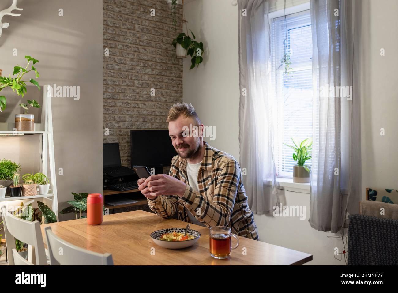 Un colpo laterale di un giovane seduto ad un tavolo nel suo soggiorno rilassante, sta usando il suo smartphone e ha un piatto di pasta vicino. Foto Stock