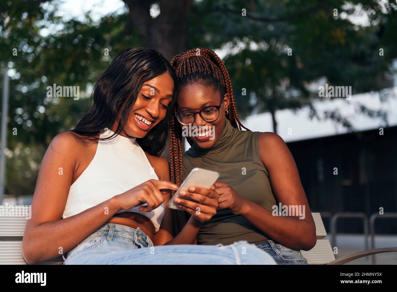 due amici neri che si divertono seduti su una panca del parco guardando un telefono cellulare, concetto di gioventù e comunicazione Foto Stock