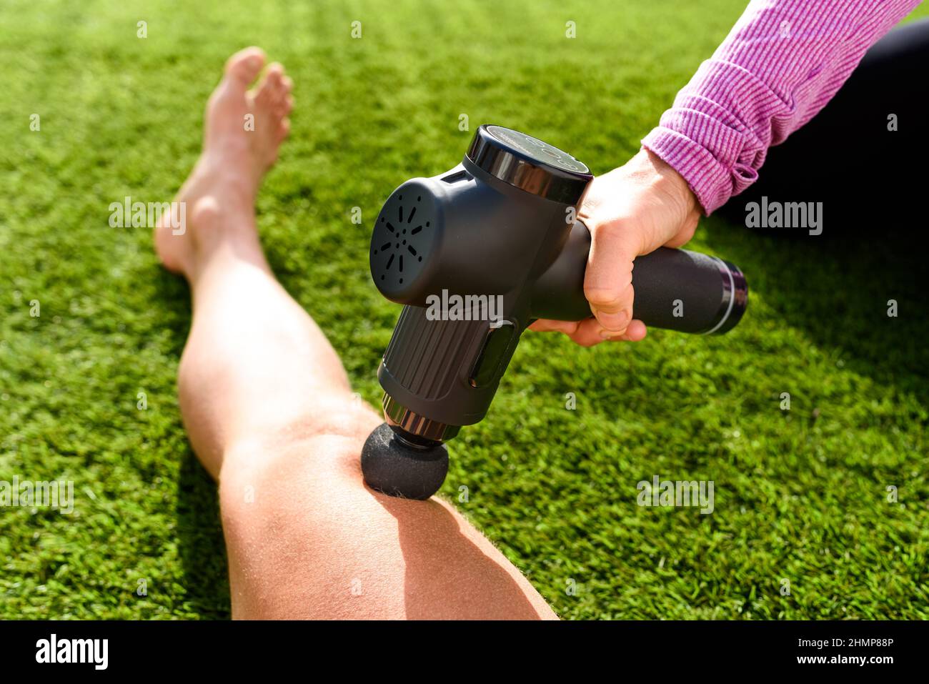 Assistente massaggia le gambe di un atleta con una pistola da massaggio a percussione per recuperare da un allenamento intenso. Foto Stock