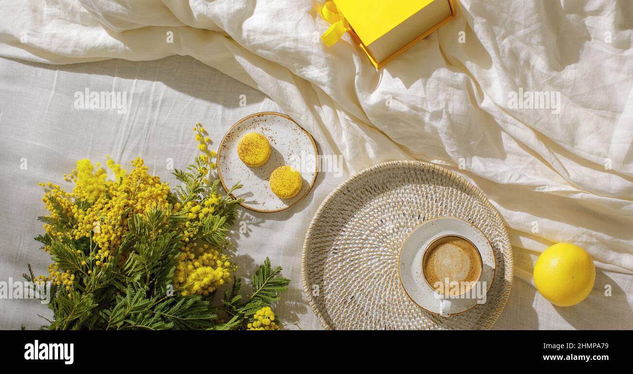 Rami di fiori gialli di mimosa su lenzuola di lino bianco, tazza o caffè, amaretti gialli francesi, pompelmo e confezione regalo gialla. Spazio copia per con Foto Stock