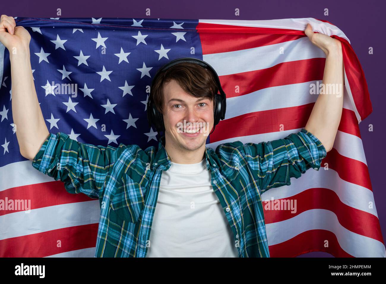 Ritratto di studente maschio felice in camicia verde indossando cuffie tenendo bandiera americana su sfondo viola sorridente e guardando la macchina fotografica Foto Stock