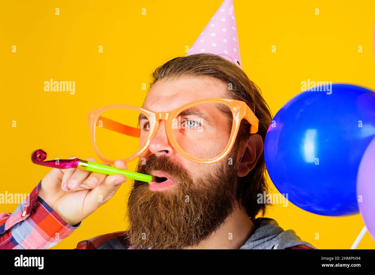 Uomo portatore in berretto di compleanno con corno partito. Maschio in cappello a cono di carta e bicchieri grandi con noisemaker. Foto Stock
