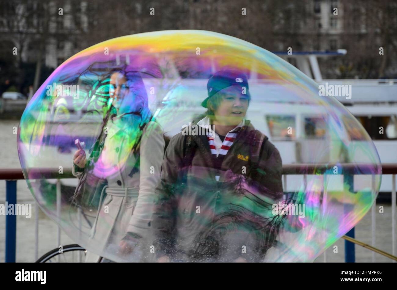 Un artista di strada sul lungofiume del Tamigi a Londra, Regno Unito, rilascia bolle. Un ragazzo e una donna che guardano attraverso una grande bolla traslucida Foto Stock