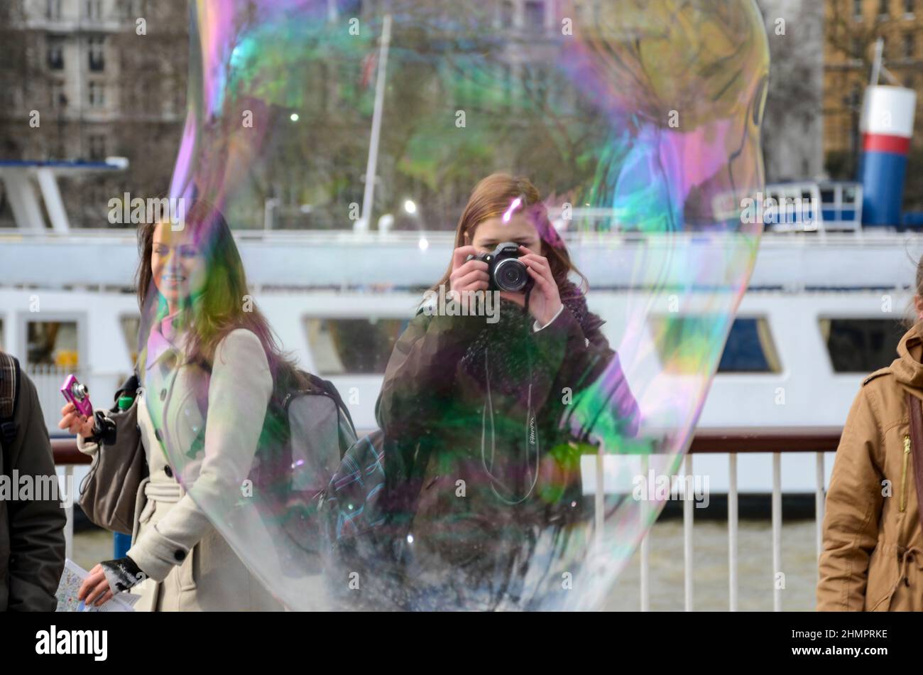 Un artista di strada sul lungofiume del Tamigi a Londra, Regno Unito, rilascia bolle. Turisti che scattano fotografie attraverso una grande bolla Foto Stock