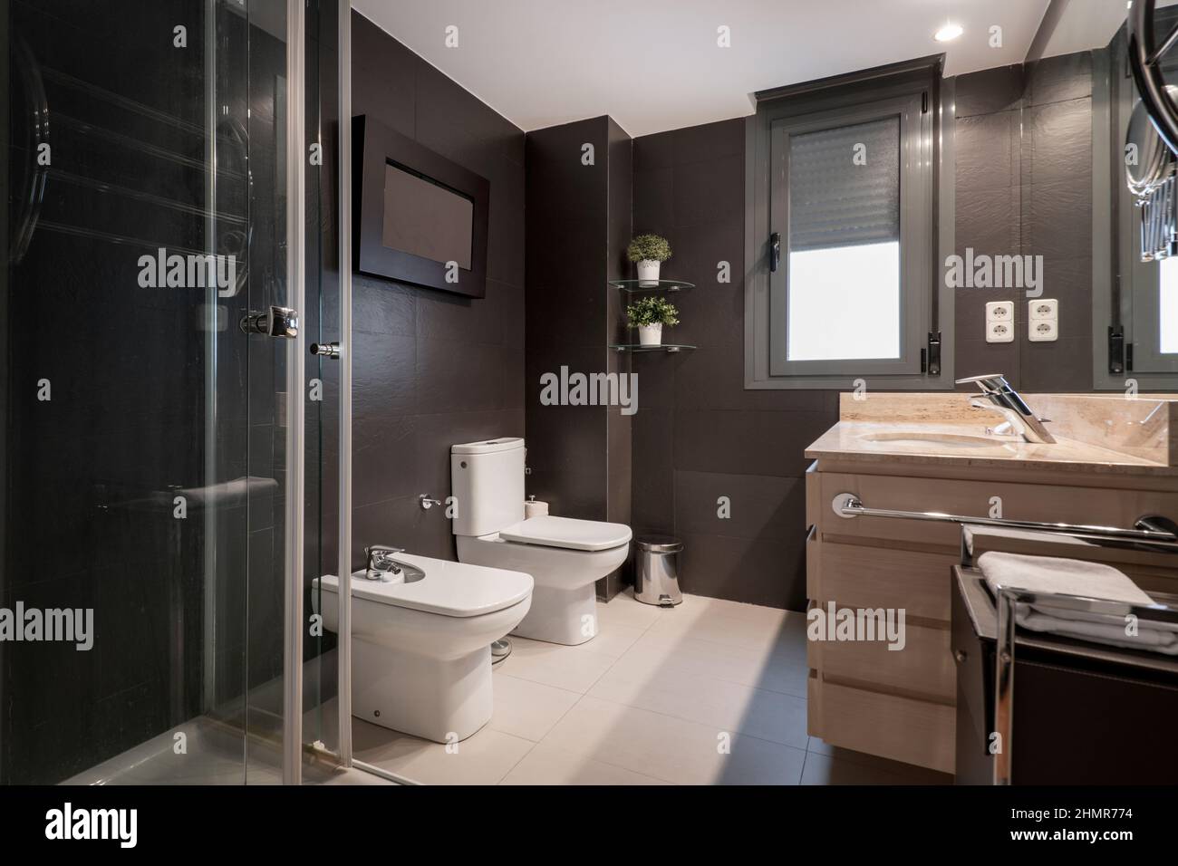 Moderno bagno con pareti piastrellate marrone scuro, doccia, toilet ciotole bianche e piano di lavoro in marmo color crema in un appartamento per vacanze in affitto Foto Stock