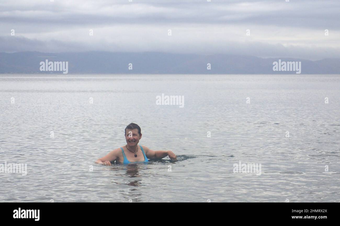 Una donna di mezza età (63) che gode di una nuotata estiva in mare sotto il cielo grigio al largo dell'isola di Kapiti, con la nuvola bassa sopra l'isola del Nord della Nuova Zelanda sullo sfondo Foto Stock