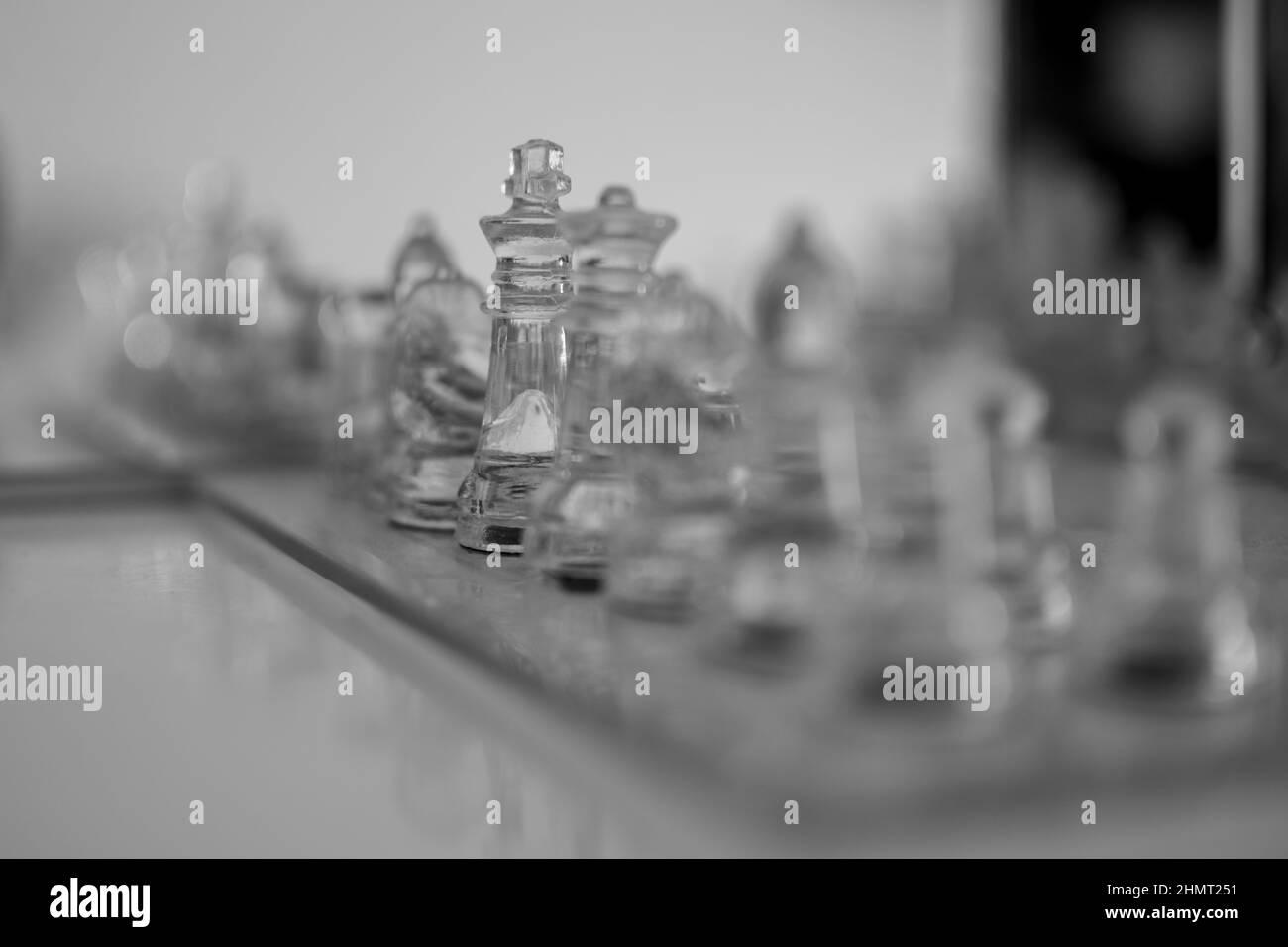 Set riflesso di pezzi di scacchi di vetro sfocato primo piano con la regina e il re focalizzato. Scala di grigi colore sfondo bianco. Foto Stock