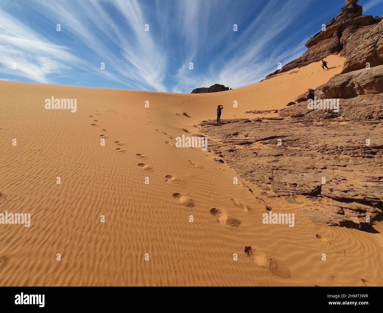 Vista ad angolo basso di un uomo che filma un altro con il suo telefono, iniziare a correre verso il basso una duna di sabbia rocciosa, impronte parallele. Cielo blu nuvoloso sfondo. Foto Stock