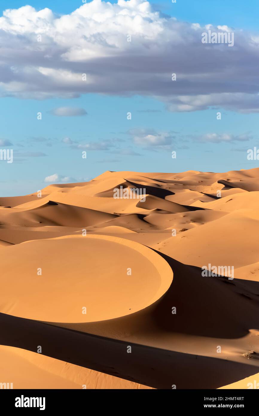 Forme contrastanti e ombre di dune di sabbia color giallo dorato e arancio e cielo blu nuvoloso nel vasto paesaggio del deserto del Sahara in Algeria. Foto Stock