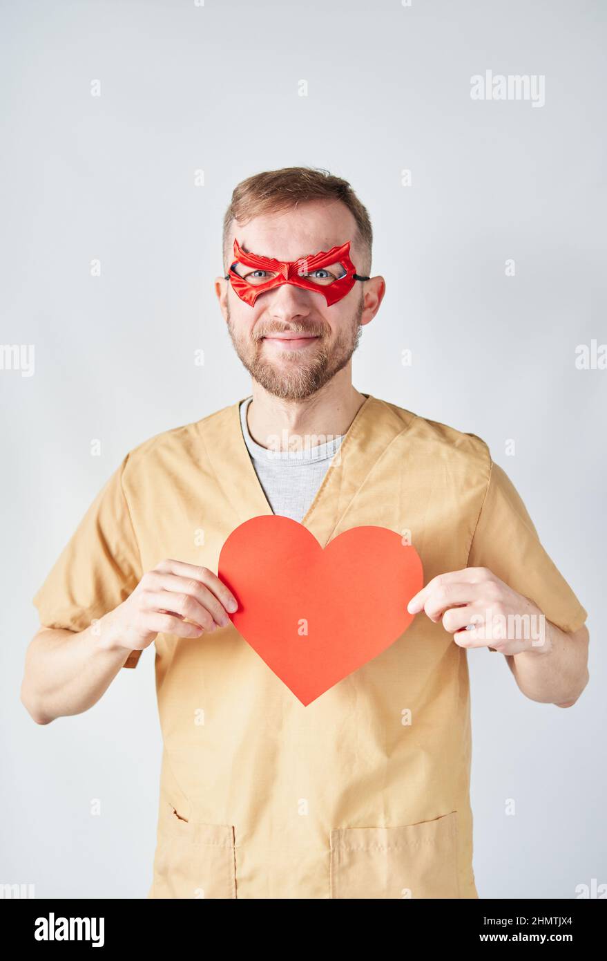 Allegro medico cardiologo maschile caucasico in uniforme medica e maschera di supereroe rosso con carta rossa a forma di cuore. Medico chirurgo adulto in ospedale che si posa sopra la parete. Foto di alta qualità Foto Stock