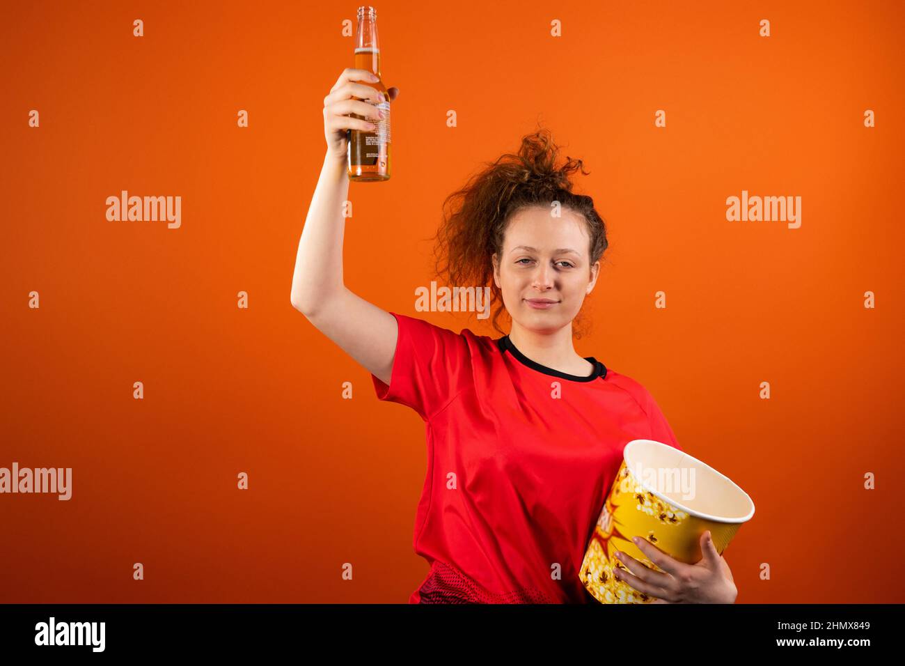 Donna di football rilassato con una divisa rossa con una ciotola di popcorn e una bottiglia di birra che solleva la mano. Foto Stock