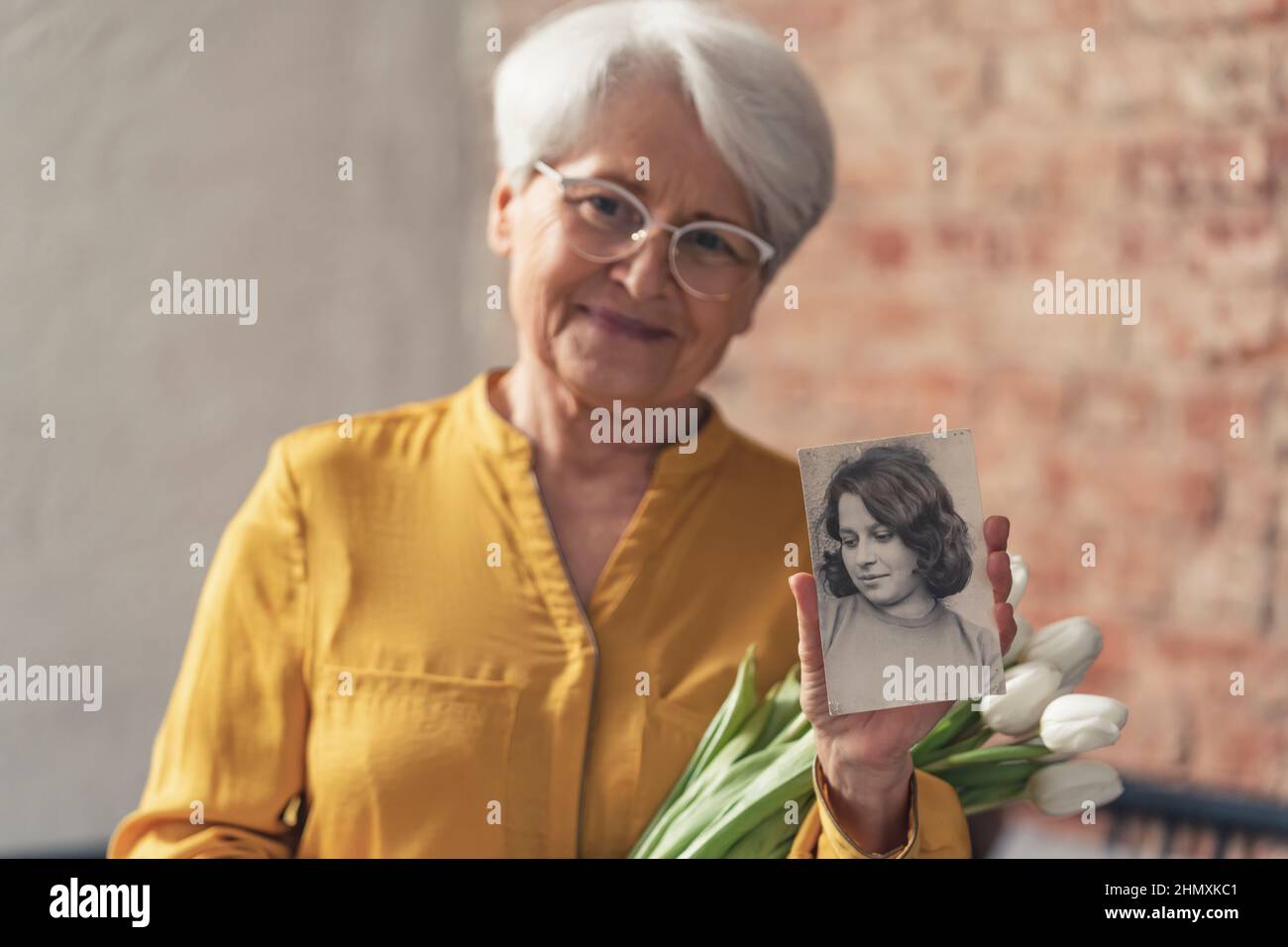 donna più anziana in pensione caucasica che tiene un mazzo di fiori bianchi e un ritratto bianco e nero dal suo passato. Foto di alta qualità Foto Stock