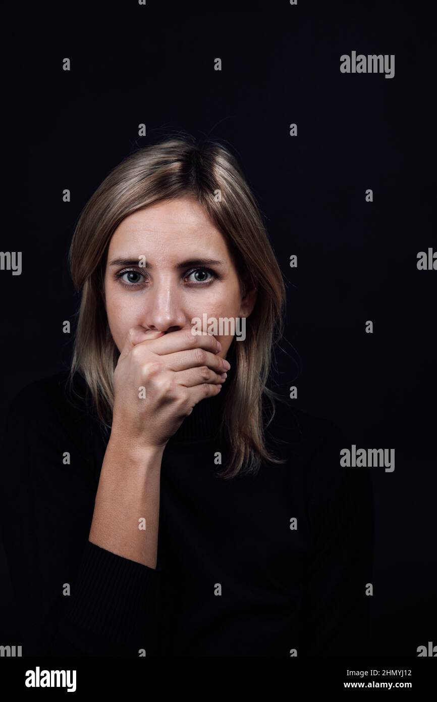 Donna minacciata con mano sulla bocca che la rende silenziosa in blusa nera su sfondo nero. Vittima di abuso fisico e psicologico. Illuminazione a gas Foto Stock
