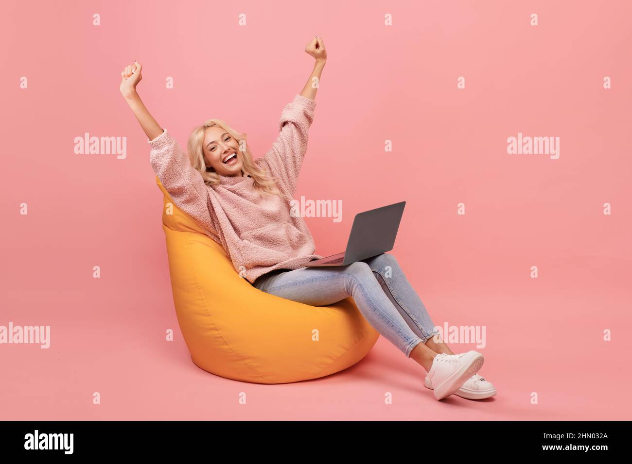 Donna allegra in casual seduta in poltrona beanbag con computer portatile e sollevare le mani, ha ottenuto il lavoro da sogno o grandi notizie Foto Stock
