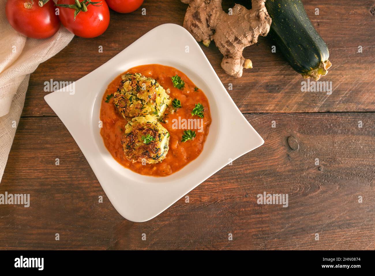 Palle di verdure fritte di zucchine e patate in salsa di pomodoro in un moderno piatto bianco e ingredienti su un rustico tavolo di legno, vegano e vegetariano Foto Stock