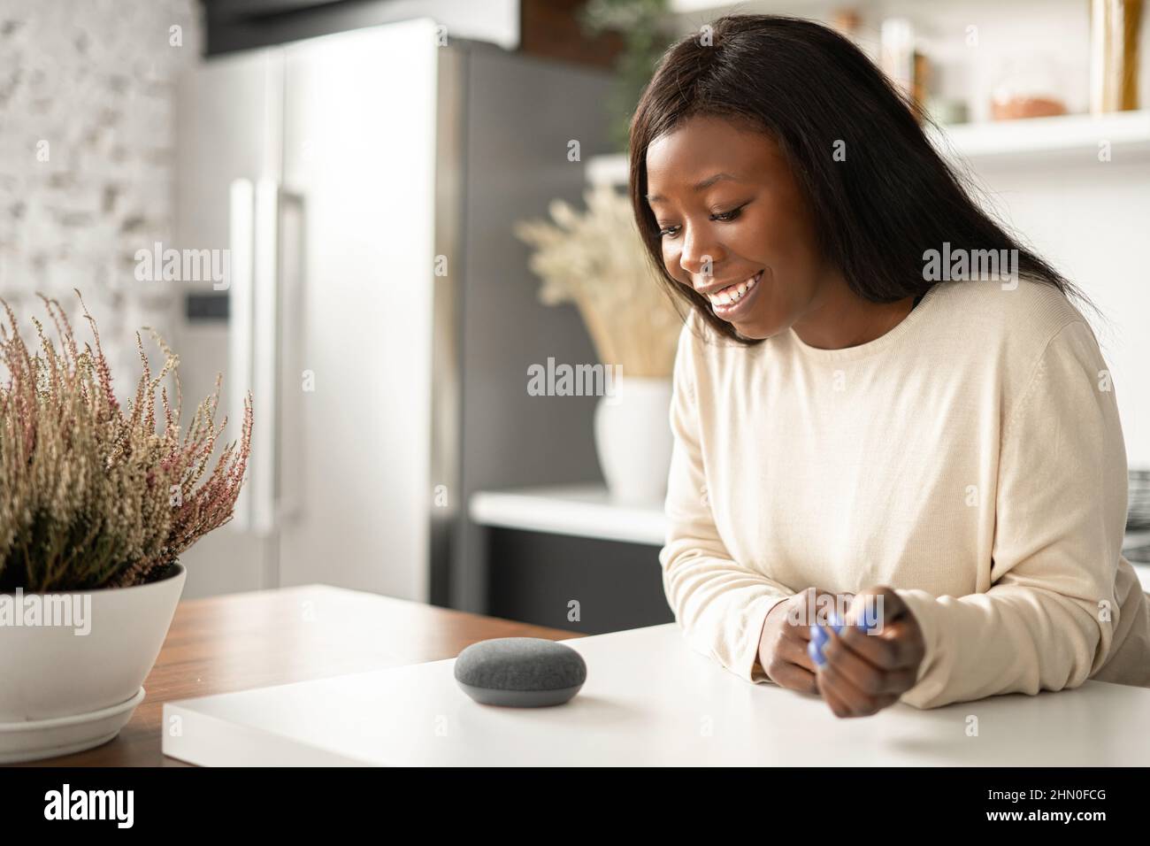 Sorridente donna afroamericana sta usando il dispositivo di assistente domestico intelligente con la funzione di riconoscimento vocale, signora multirazziale che parla con l'altoparlante intelligente portatile. Concetto di casa intelligente Foto Stock