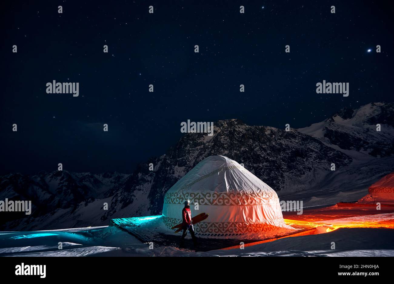 Uomo a piedi con snowboard spaccato vicino Yurt casa nomade presso la stazione sciistica di Shymbulak in Almaty, Kazakistan. Astrofotografia notturna invernale con stelle e. Foto Stock