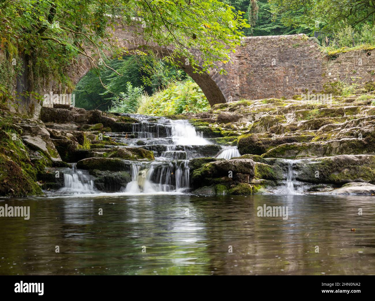 Vista estiva di Walden Beck a West Burton, Yorkshire Dales, con il torrente che passa sotto un ponte di cavalli da imballaggio e cascata su gradini di pietra calcarea Foto Stock