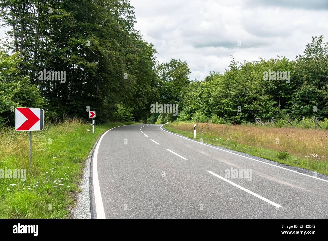 Chevron strada segnaletica su una curva stretta lungo una strada di campagna in una nuvolosa giornata estiva Foto Stock Chevron strada segnaletica su una curva stretta lungo una strada di campagna in una nuvolosa giornata estiva Foto Stock