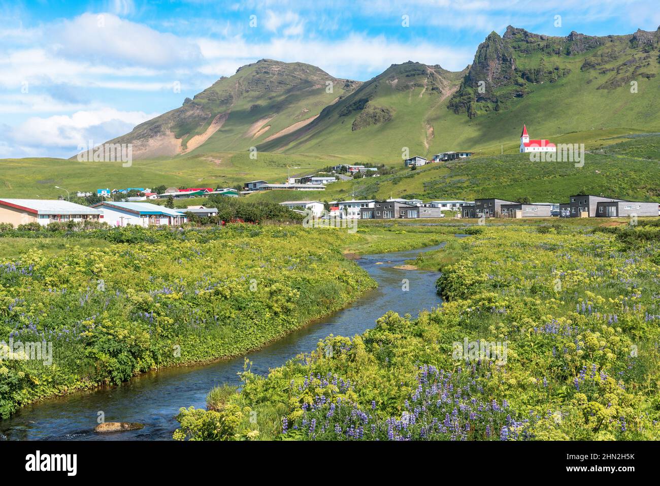 Piccola città dominata da una piccola chiesa con un tetto rosso in una giornata estiva soleggiata. Sullo sfondo vi sono delle torreggianti montagne verdi. Foto Stock